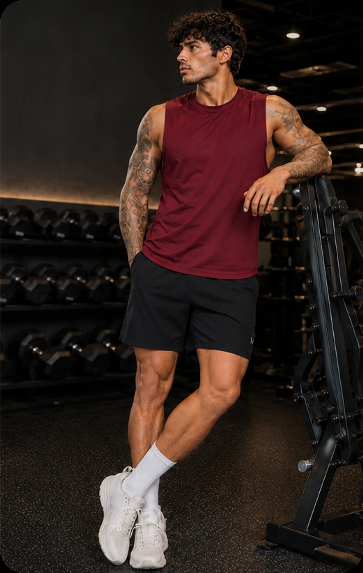 Model in a maroon cut-off tank and black training shorts by the dumbbell rack.