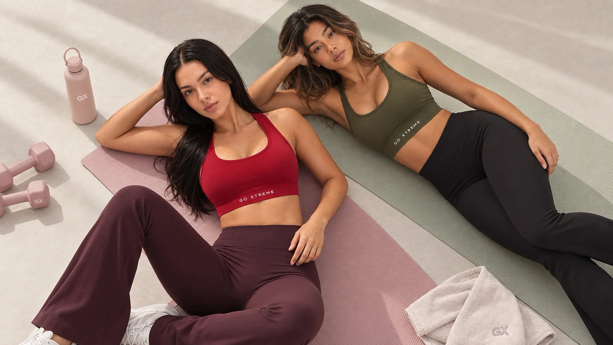 Two models in GX sports bras and leggings, crimson and olive, resting on training mats in the studio.
