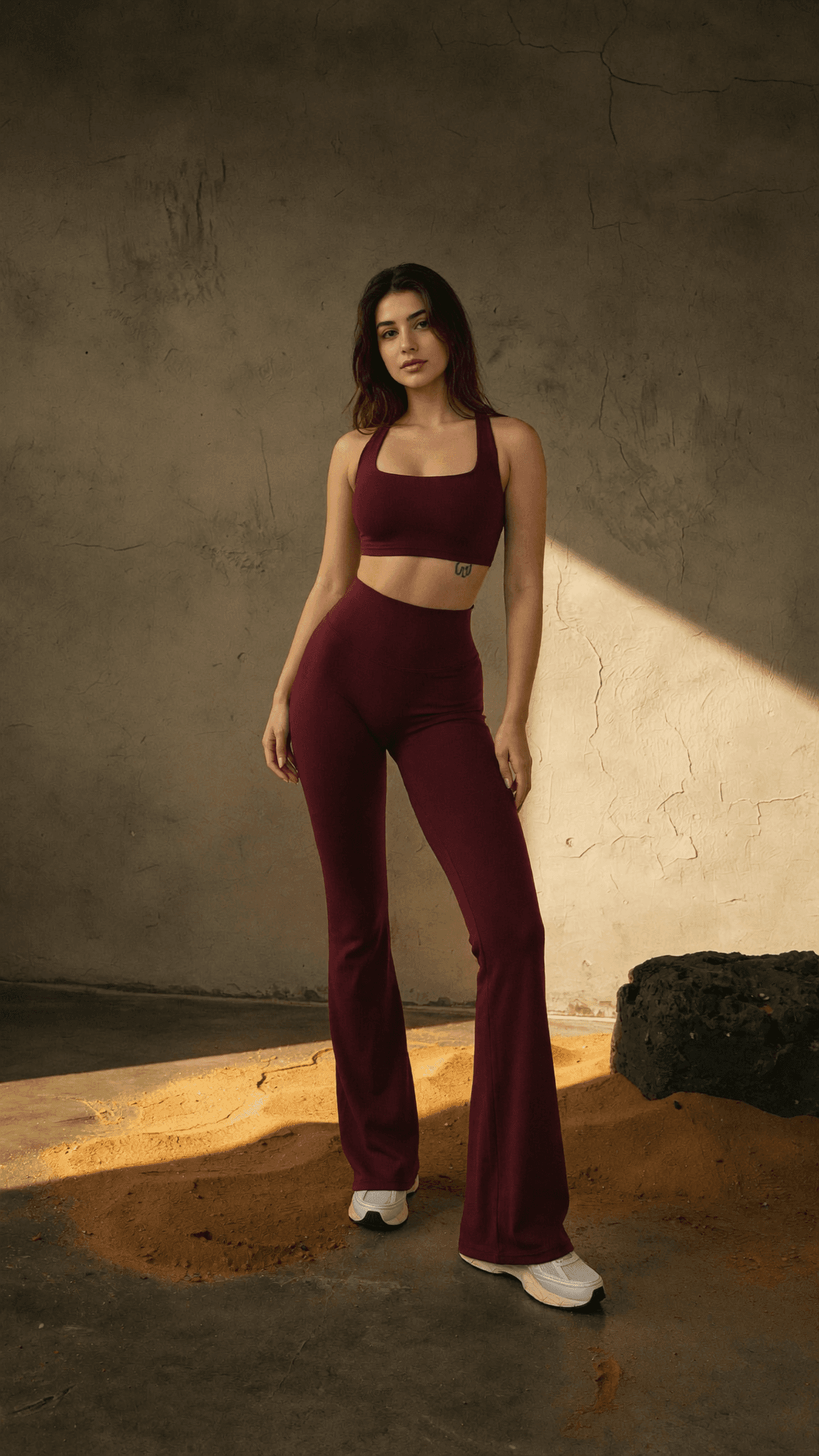 A model in the maroon GX sports bra and flared leggings, standing in warm light against a plaster wall